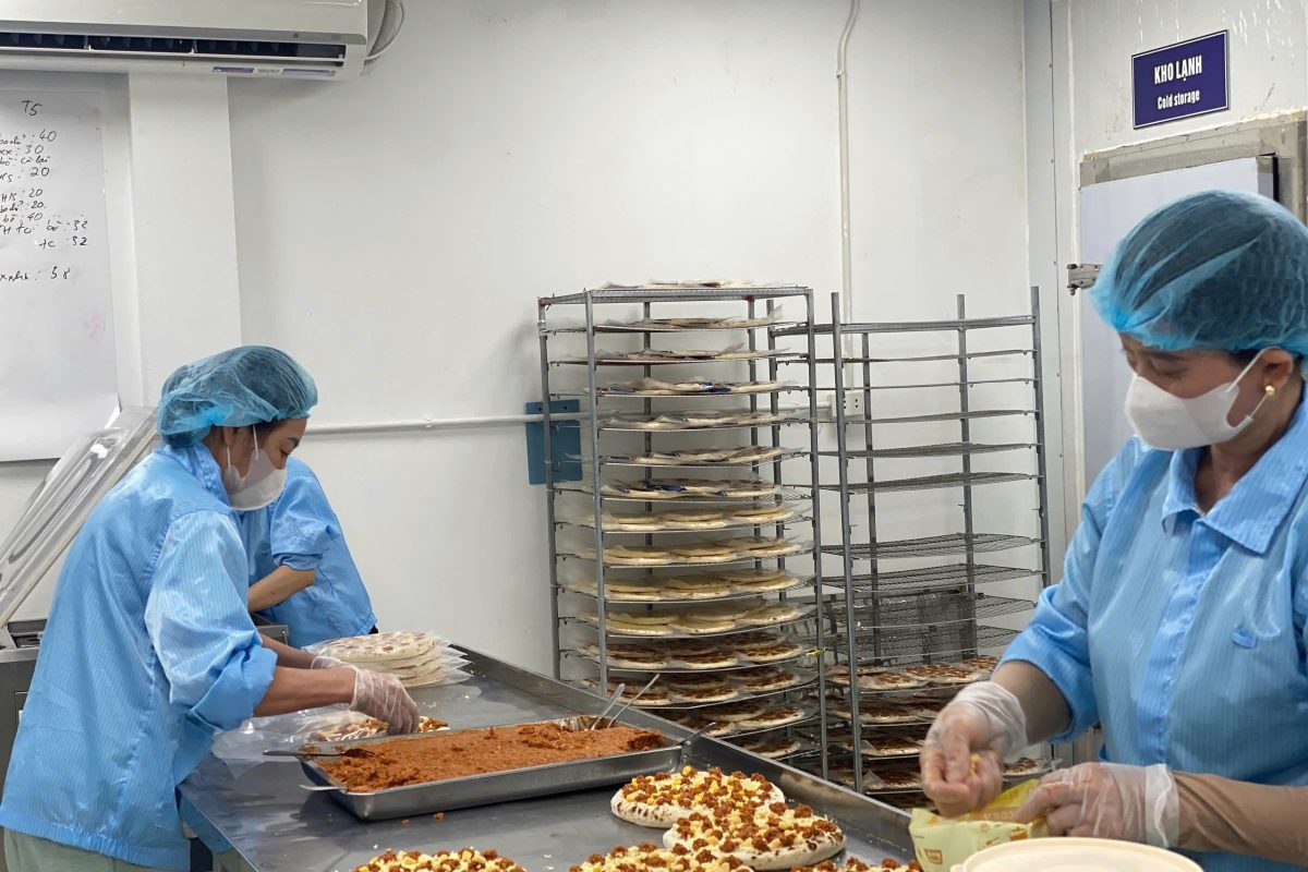 Nhân viên FoodTek phủ nhân pizza trong phòng sạch, bên cạnh cửa kho lạnh của xưởng